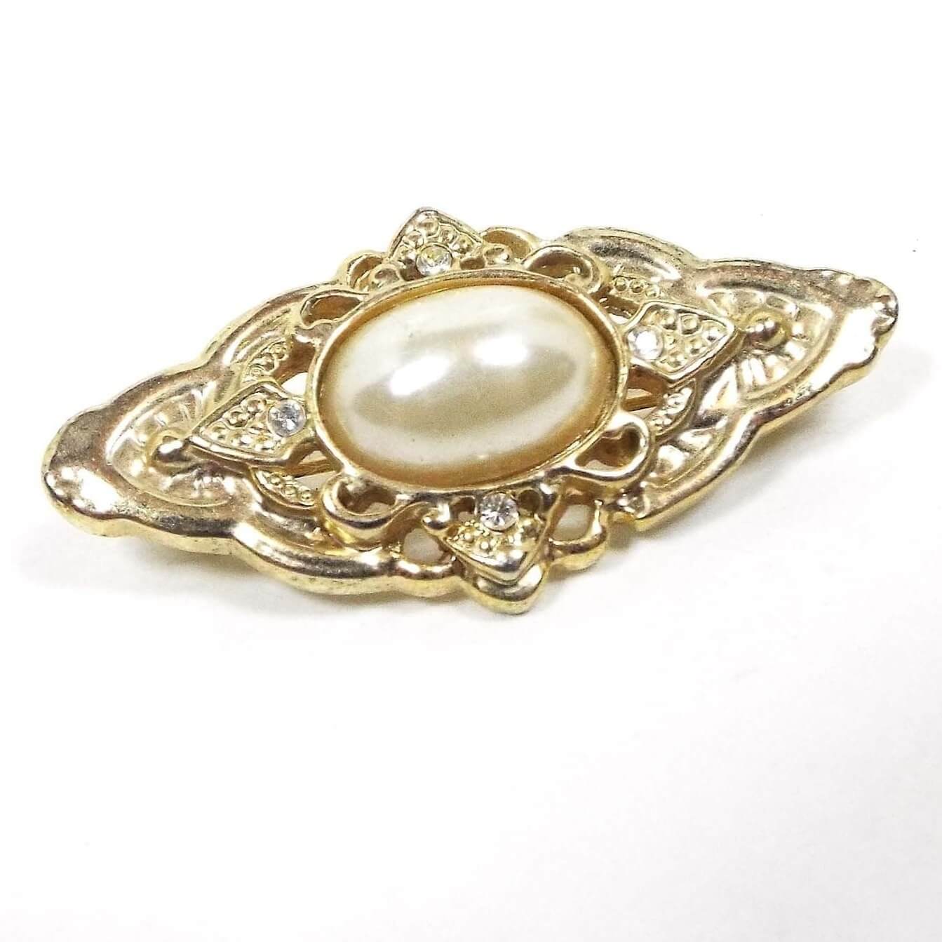 Front view of the retro vintage faux pearl brooch pin. The brooch metal is gold tone in color and has a large marquis like shape. There is a domed oval plastic imitation pearl in the middle. The rest of the brooch has a cut out filigree style design of differing shapes and patterns.
