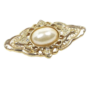 Front view of the retro vintage faux pearl brooch pin. The brooch metal is gold tone in color and has a large marquis like shape. There is a domed oval plastic imitation pearl in the middle. The rest of the brooch has a cut out filigree style design of differing shapes and patterns.