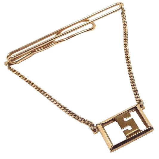 Angled front view of the 1930's vintage Swank initial tie chain bar. The metal is gold tone in color. The top has a long open oval wire style bar that curves around to the back with a wider open oval at the end. There is thin curb chain hanging from each end down to an open rectangle charm that has a block letter style initial S in the middle. 