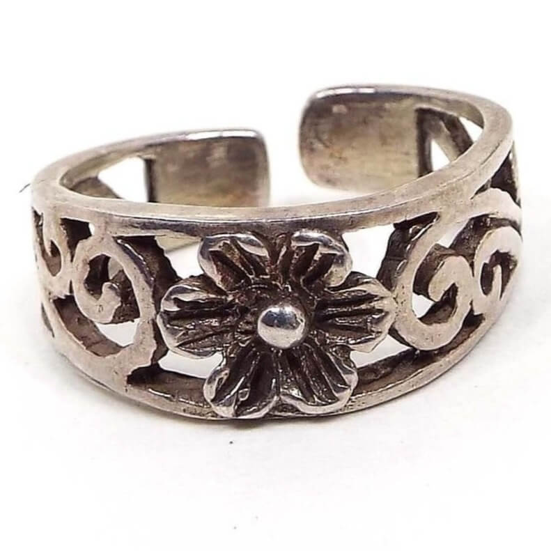 Front view of the retro vintage Boho flower toe ring. The metal is slightly darkened silver tone in color. There is a filifree swirl design around most the the ring. In the middle is a flower with textured petals. The back has rounded off ends and is open so it can be adjusted for size.