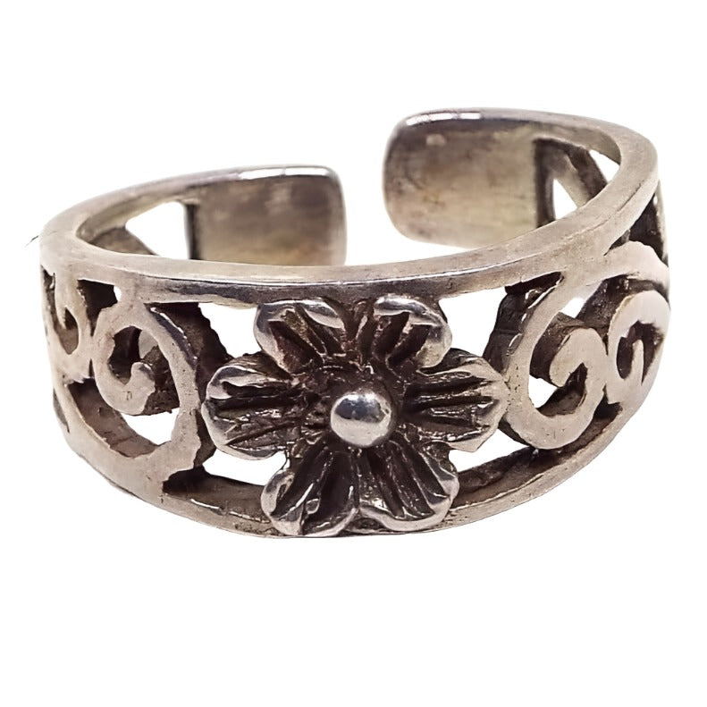 Front view of the retro vintage Boho flower toe ring. The metal is slightly darkened silver tone in color. There is a filifree swirl design around most the the ring. In the middle is a flower with textured petals. The back has rounded off ends and is open so it can be adjusted for size.