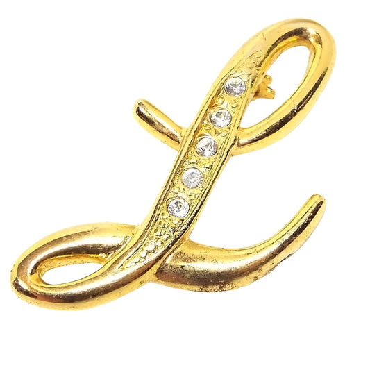 Front view of the 1980's retro vintage rhinesetone initial brooch pin. The metal is gold tone in color. It is shaped like a cursive script letter initial L and has a row of small round rhinestones through the middle part of the L. 