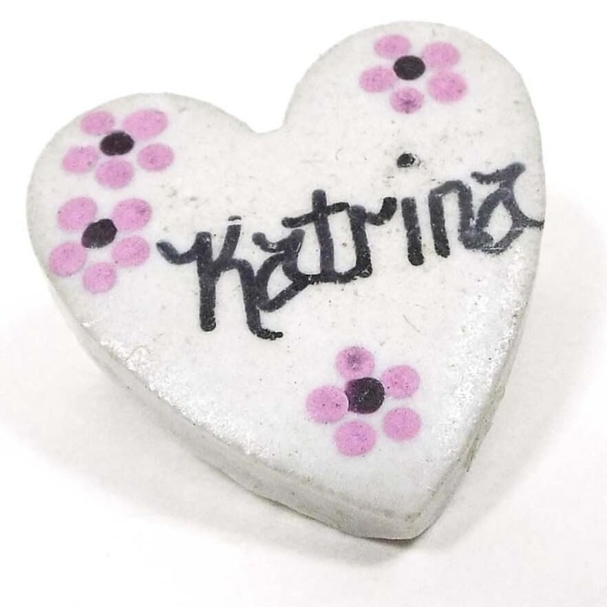 Front view of the retro vintage porcelain heart name brooch. It is white matte porcelain heart shape. In the middle of the brooch the name Katrina is painted in black fancy block style letters. There are four small painted flowers with two in one corner, one in another corner, and one at the bottom of the heart. They are painted pink with purple middles. 