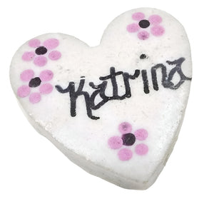 Front view of the retro vintage porcelain heart name brooch. It is white matte porcelain heart shape. In the middle of the brooch the name Katrina is painted in black fancy block style letters. There are four small painted flowers with two in one corner, one in another corner, and one at the bottom of the heart. They are painted pink with purple middles. 
