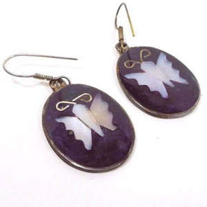Front side of the Mexico Alpaca earrings. The metal is darker silver in color from age. They are shaped like large ovals with a butterfly design in the middle. The butterfly wings and body are inlaid mother of pearl shell and are pearly white in color with hints of pin. The rest of the oval is a dark pearly purple resin enamel. Top of earrings have fish hook style pierced ear wires. 