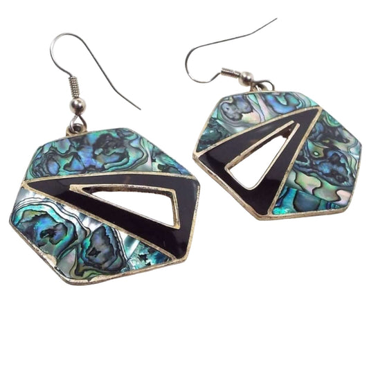 Front side of the Mexican Alpaca earrings. Metal part is silver in color. These geometric style earrings have large hexagon shaped drops that have a triangle angled in the middle with a cut out triangle area in the middle of them. The triangles have black resin enamel and the rest of the earrings are inlaid pearly multi color abalone shell. There are swirls of green, purple, blue, and gray. Tops have pierced fish hook style ear wires.