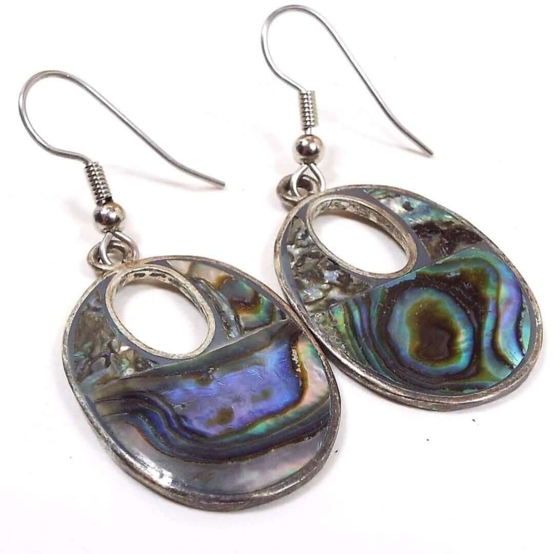 Front view of the retro vintage Mexican alpaca earrings with inlaid abalone shell. The metal is a slightly darker silver tone in color. The drops are oval shaped with a small oval cut out towards the top of them. They have pearly multi color abalone shell on the fronts.