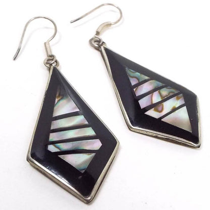 Front view of the Southwestern vintage earrings. They are angled teardrop shaped The middle area has inlaid pearly multi color abalone shell pieces in a diamond like shape with lines running between the pieces. The lines and outer edge are black resin enamel. The metal is silver tone in color and the earrings have curved wire style earwires.