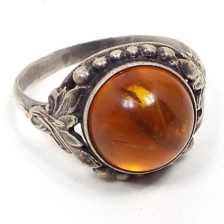 Angled front and side view of the retro vintage RJ sterling silver amber ring. The top of the ring has a domed round amber cab with swirled shades of orange. There is a sterling dot pattern on the top and bottom of the bezel area and a textured leaf like design on either side. The sterling has darkened some from age for a silver gray color.