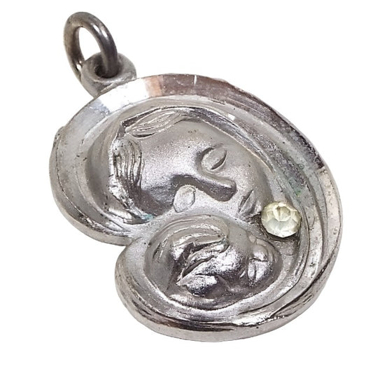 Front view of the Mid Century vintage Christian charm pendant. The metal is matte silver tone in color with a raised design of the head of Mother Mary and Baby Jesus. It has faceted etched edges and a small rhinestone just under Mary's head. There is a small ring at the top with a jump ring attached to it.