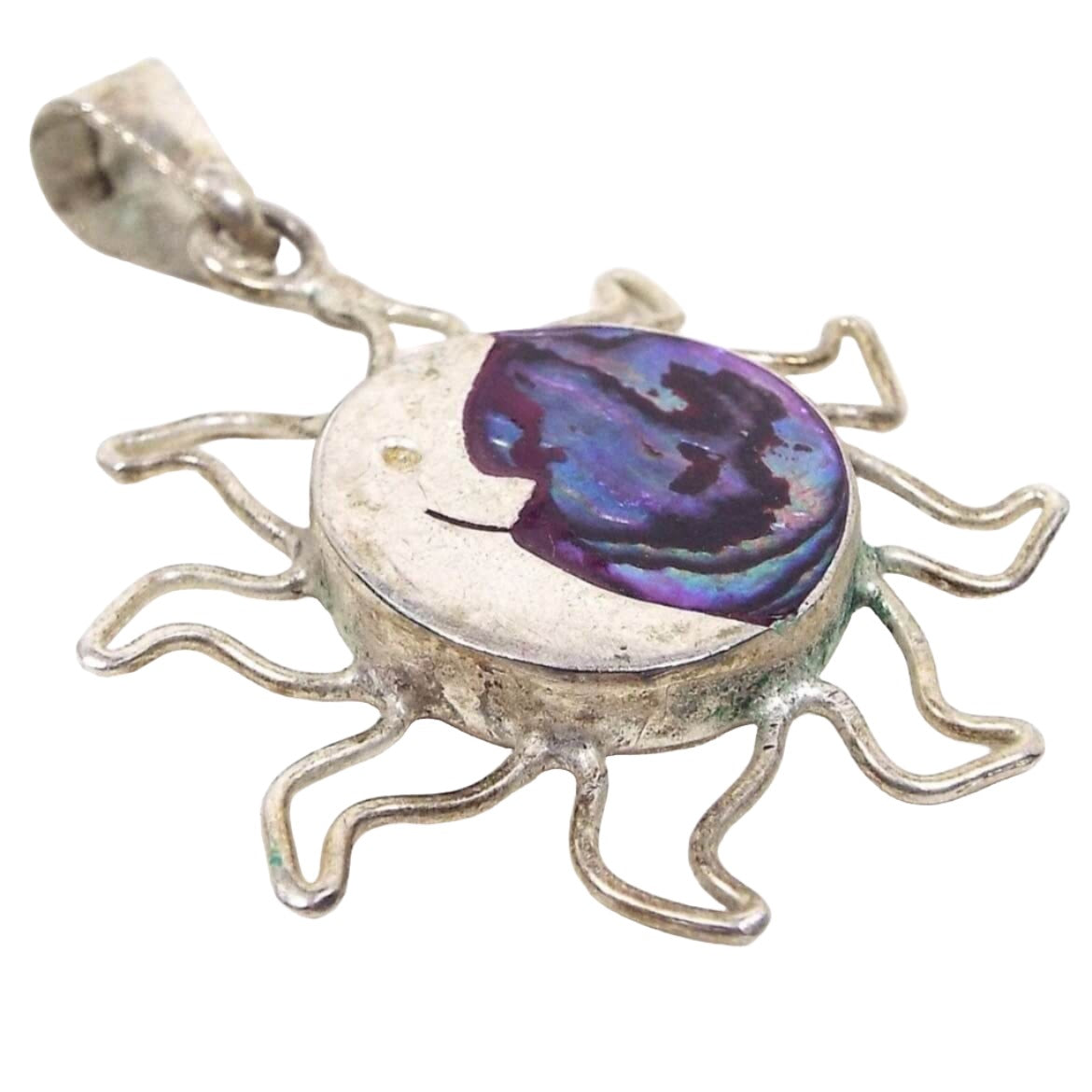 Front view of the retro vintage Mexican sterling pendant. The sterling is slightly darkened from age. The main area in the middle is round with a bezel and has open wire style flames to represent the sun part of the pendant. The inside of the circle has a moon shape on the left with a dot for an eye and a half smile and the other side has inlaid abalone shell. The abalone shell has marbled and swirled colors in blues and purples that have flashes of color as you move around. There is a bail at the top.
