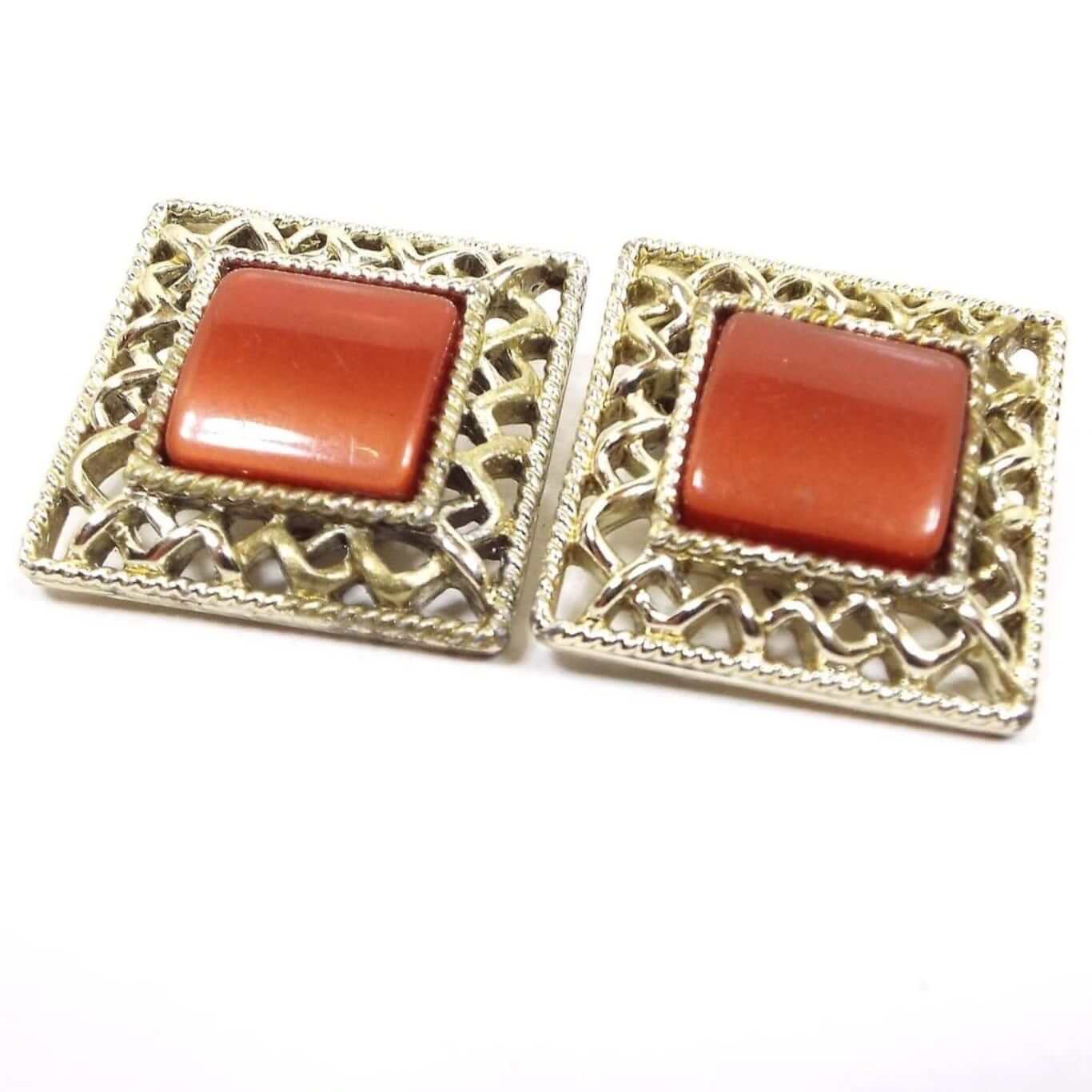 Front view of the Mid Century vintage Coro clip on earrings. They are square in shape with puffy middle lucite plastic cabs. The cabs are a pearly burnt orange in color and are surrounded by a gold tone color woven style filigree edge. 