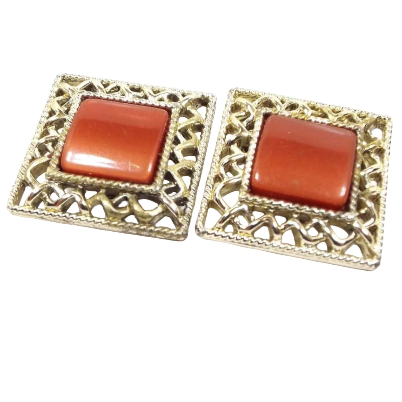 Front view of the Mid Century vintage Coro clip on earrings. They are square in shape with puffy middle lucite plastic cabs. The cabs are a pearly burnt orange in color and are surrounded by a gold tone color woven style filigree edge. 