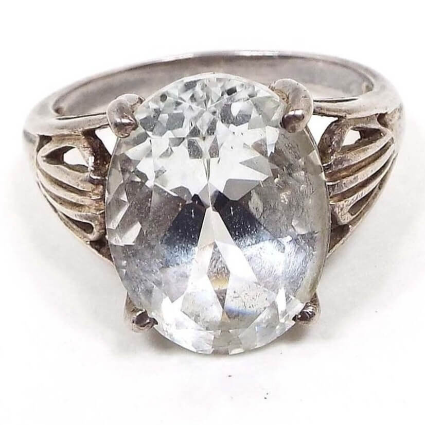 Angled top view of the retro vintage SCBS sterling silver white topaz solitaire statement ring. The top has a large prong set oval white topaz that is almost clear in color. The sterling band flares a the top and has a split band design with a cut out pattern in the middle. 